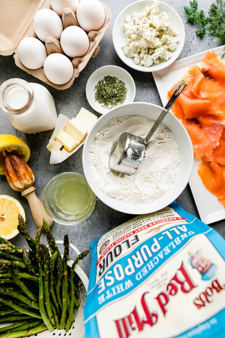 Savory dutch baby ingredients arranged on a light gray surface: unsalted butter, Bob's Red Mill Unbleached White All-Purpose Flour, whole milk, large eggs, dried thyme, fresh asparagus, lemon, smoked salmon, Boursin cheese, fresh herbs, kosher salt, and ground black pepper.