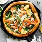 An assembled herby dutch baby with smoked salmon, asparagus, and a Boursin cheese fills a large black cast iron skillet. The Dutch baby is garnished with lemon wedges, a lemon butter sauce, and fresh herbs. The skillet sits atop a light gray textured surface surrounded by bowls of toppings and a small glass bowl filled with lemon butter sauce. A light cream linen napkin is tied around the skillet handle and a serving spatula rests alongside.