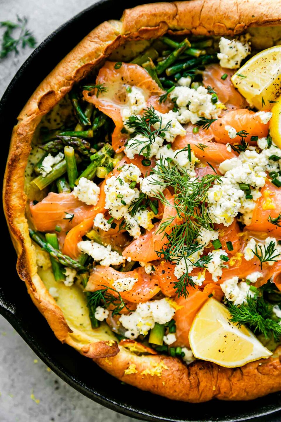 A close up macro shot of a savory dutch baby topped with smoked salmon, asparagus, and a Boursin cheese fills a large black cast iron skillet. The Dutch baby is garnished with lemon wedges, a lemon butter sauce, and fresh herbs. The skillet sits atop a light gray textured surface.
