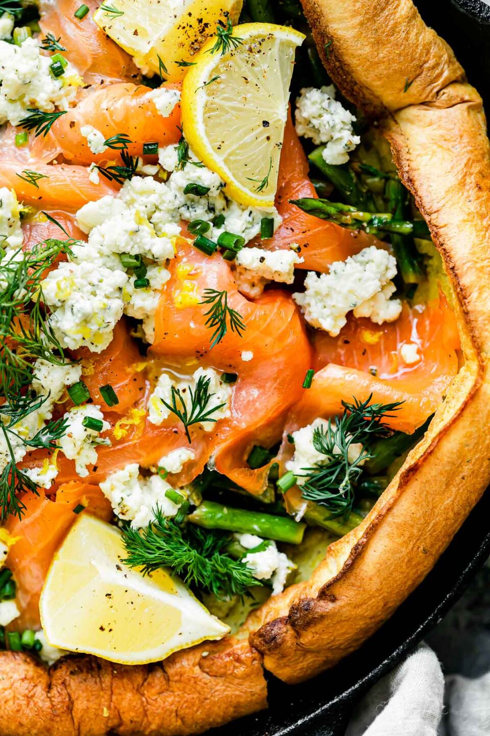 A macro close up shot of a savory dutch baby topped with smoked salmon, asparagus, and a Boursin cheese fills a large black cast iron skillet.