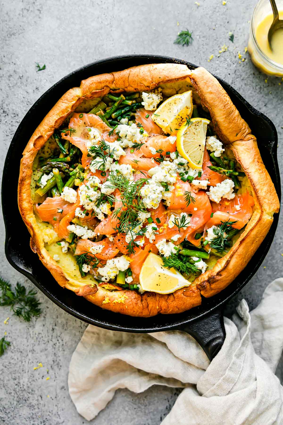 An assembled Dutch baby topped with smoked salmon, asparagus, and a Boursin cheese fills a large black cast iron skillet. The Dutch baby is garnished with lemon wedges, a lemon butter sauce, and fresh herbs. The skillet sits atop a light gray textured surface with a light cream linen napkin tied around the skillet handle. A lemon butter sauce fills a small glass bowl & rests alongside the skillet at center.
