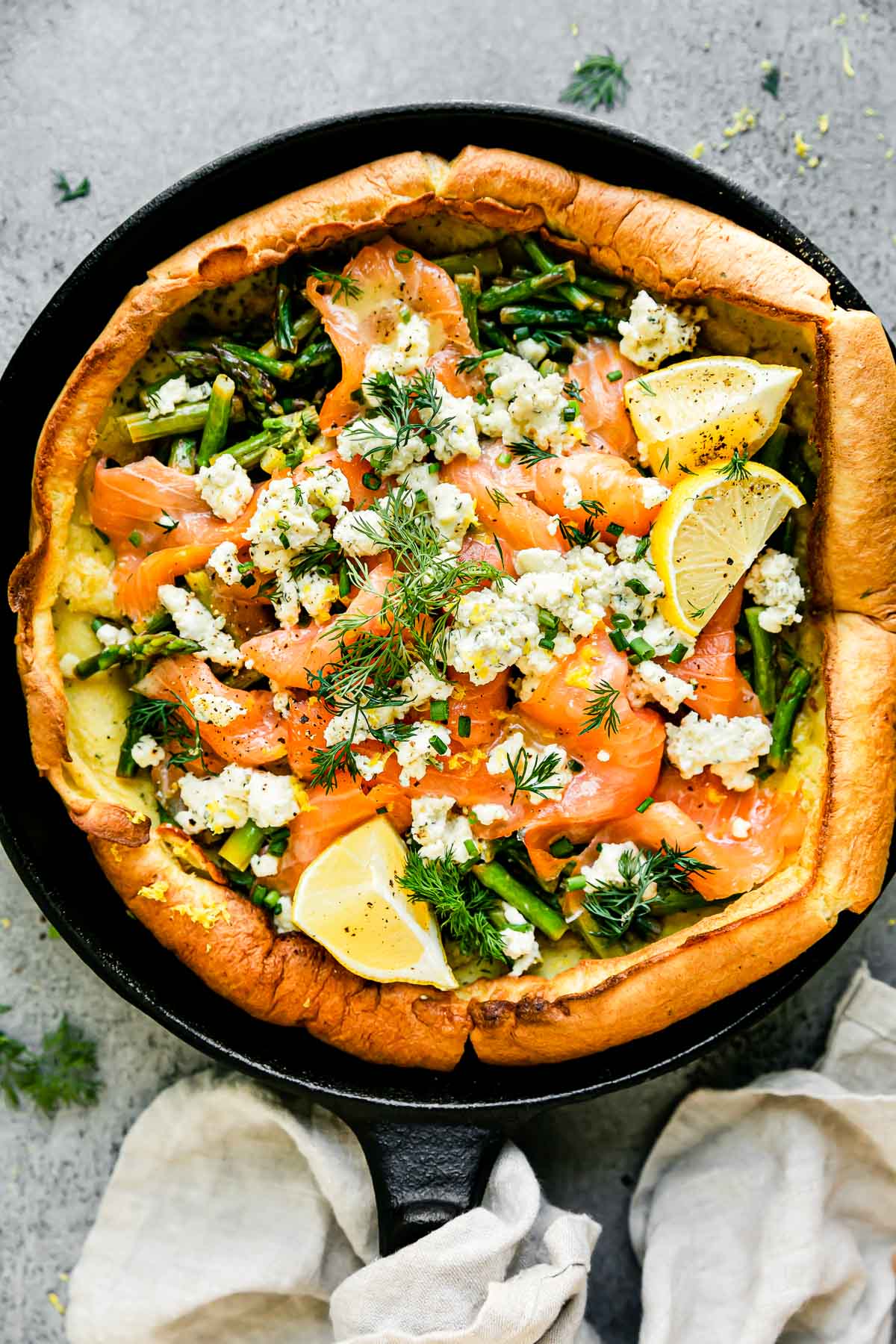 An assembled Dutch baby topped with smoked salmon, asparagus, and a Boursin cheese fills a large black cast iron skillet. The Dutch baby is garnished with lemon wedges, a lemon butter sauce, and fresh herbs. The skillet sits atop a light gray textured surface with a light cream linen napkin tied around the skillet handle.