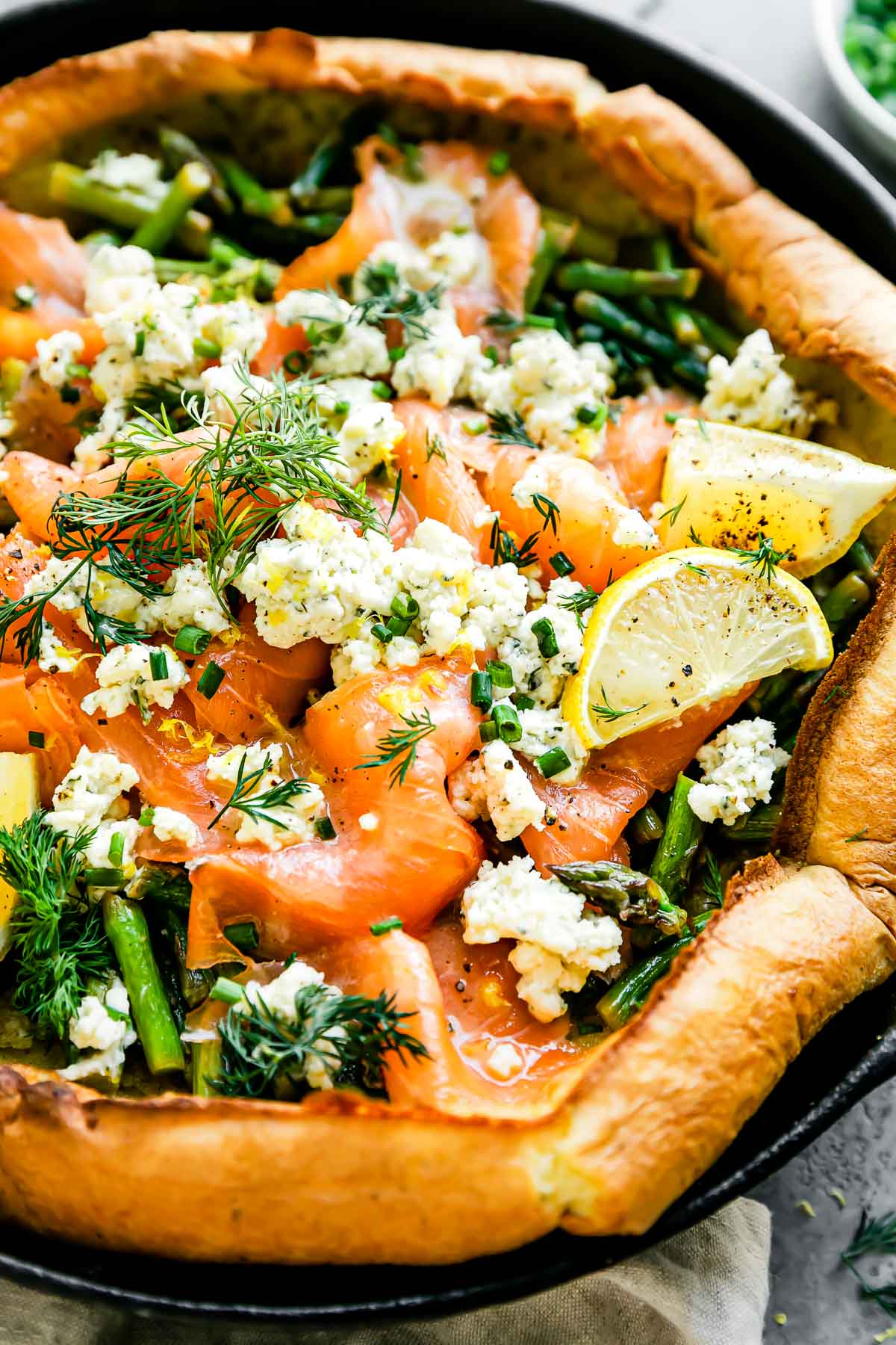 A macro close up and slightly side angle shot of an assembled Dutch baby topped with smoked salmon, asparagus, and a Boursin cheese fills a large black cast iron skillet. The Dutch baby is garnished with lemon wedges, a lemon butter sauce, and fresh herbs. The skillet sits atop a light gray textured surface with a light cream linen napkin tied around the skillet handle.