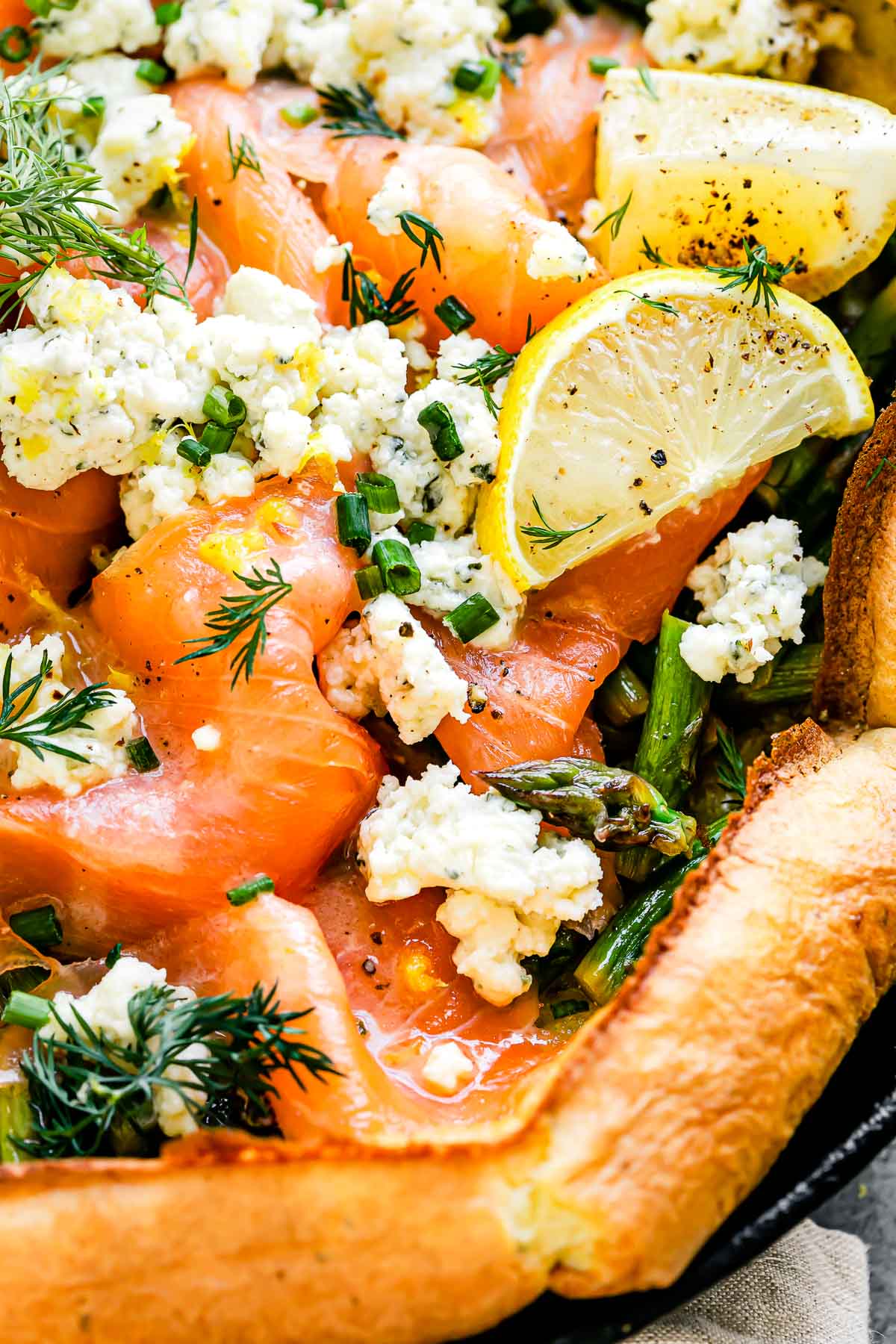 A close up macro shot of an assembled Dutch baby topped with smoked salmon, asparagus, and a Boursin cheese fills a large black cast iron skillet. The Dutch baby is garnished with lemon wedges, a lemon butter sauce, and fresh herbs.