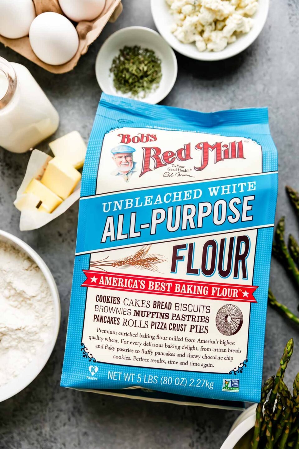 Bob's Red Mill Unbleached White All-Purpose Flour lays label side facing up atop a light gray textured surface surrounded by savory dutch baby ingredients: whole milk, eggs, Boursin cheese, unsalted butter, dried thyme, fresh asparagus, and a bowl of flour.