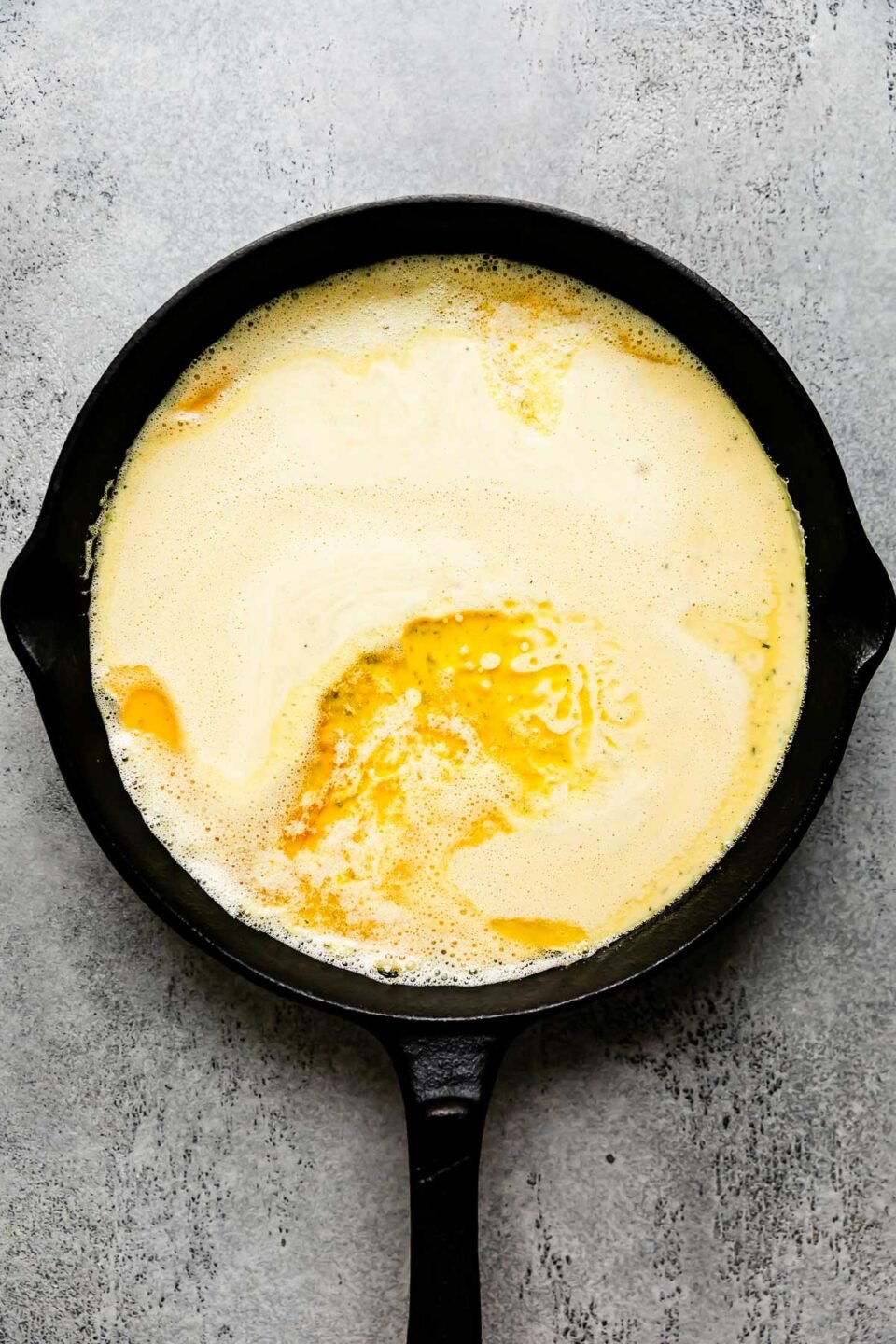 Savory dutch baby batter is poured into a large preheated cast iron skillet that sits atop a light gray textured surface.