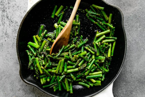1/2-inch pieces of asparagus cook inside of a large white Staub cast iron skillet that sits atop a light gray textured surface.