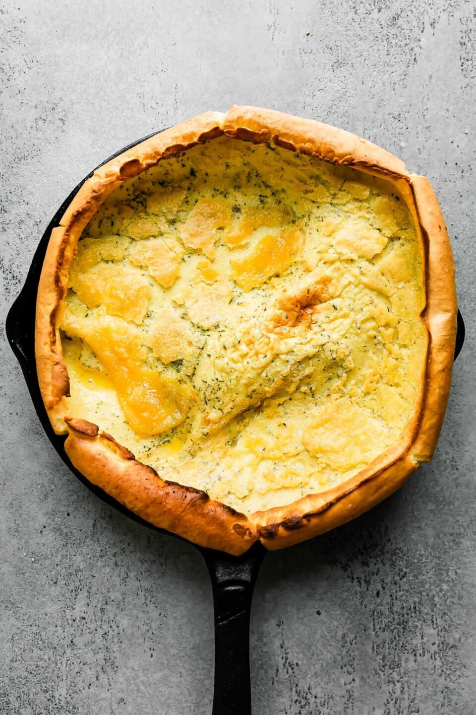 A baked savory dutch baby fills a large black cast iron skillet that sits atop a light gray textured surface.