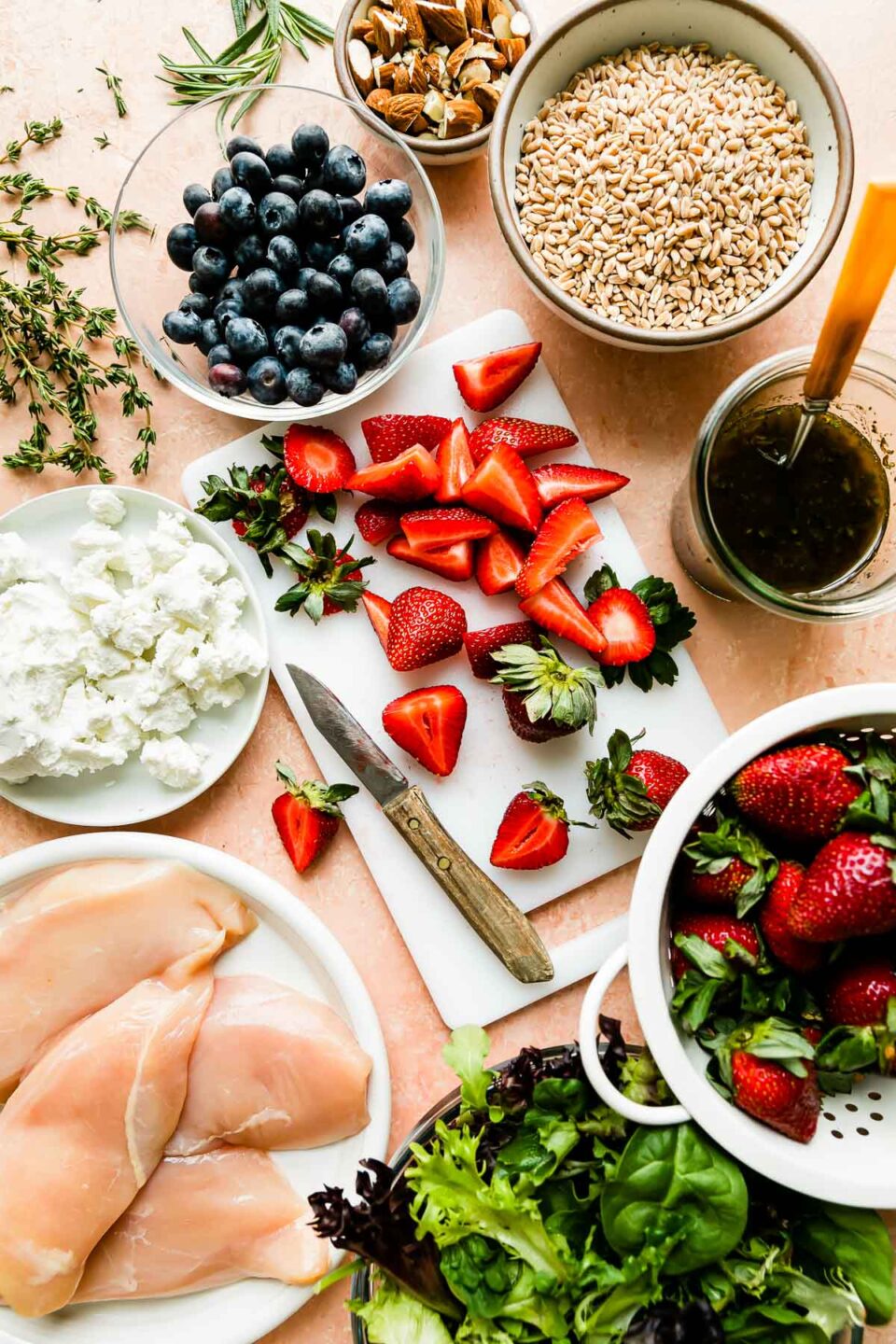 An overhead shot of Strawberry Chicken Salad ingredients are displayed on a pale peach marbled surface: strawberries, blueberries, chicken breasts, spring greens, farro, maple balsamic vinaigrette, goat cheese, almonds and fresh herbs. 