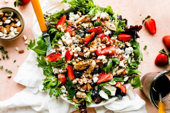 An overhead shot of the strawberry chicken salad on a light pink surface. The bowl is surrounded by a white dish towel, a jar of vinaigrette, and small bowls of strawberries, almonds and goat cheese.