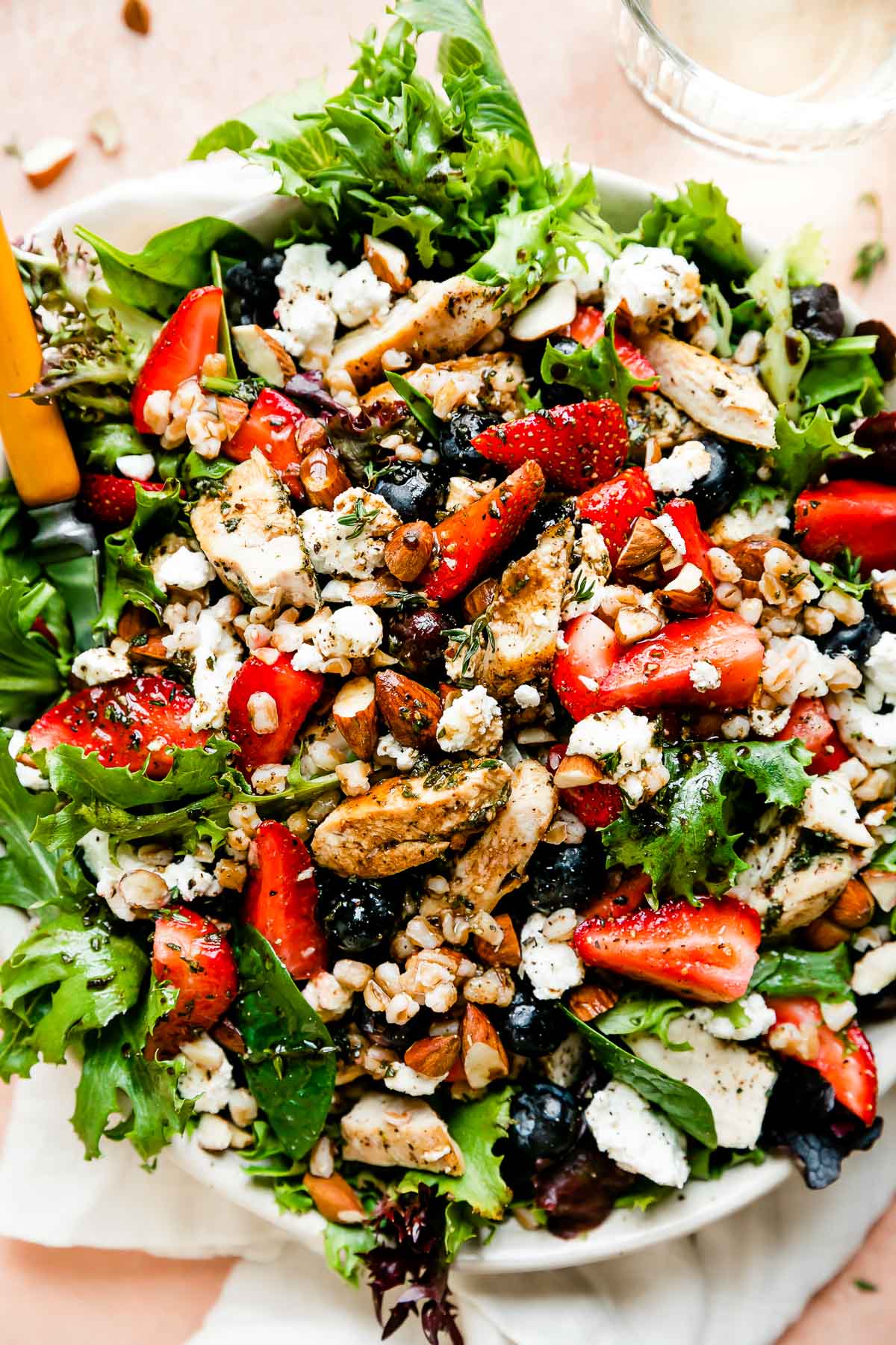 An overhead close-up shot of the strawberry chicken salad in a bowl atop a light pink surface: greens, strawberries, chicken, almonds, goat cheese and vinaigrette. A wood-handled fork is resting in the bowl and a glass of water sits alongside it.