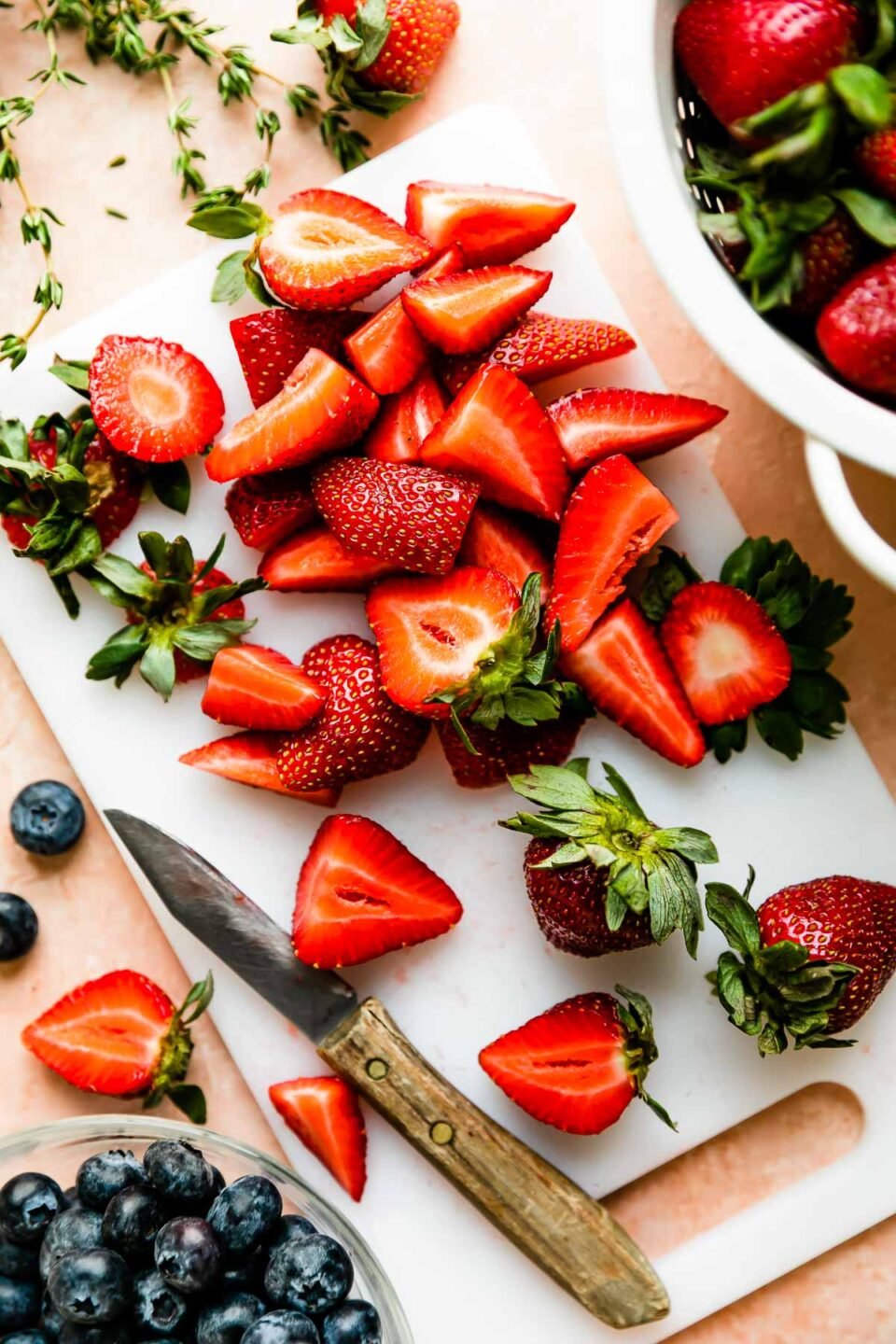Overhead shot of sliced strawberries on a white cutting board resting on a light pink marbled surface. A bowl of blueberries and a colander of strawberries sit alongside it.