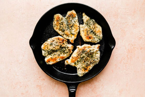 A cast-iron skillet with browned chicken breasts rests atop a pink marbled surface.