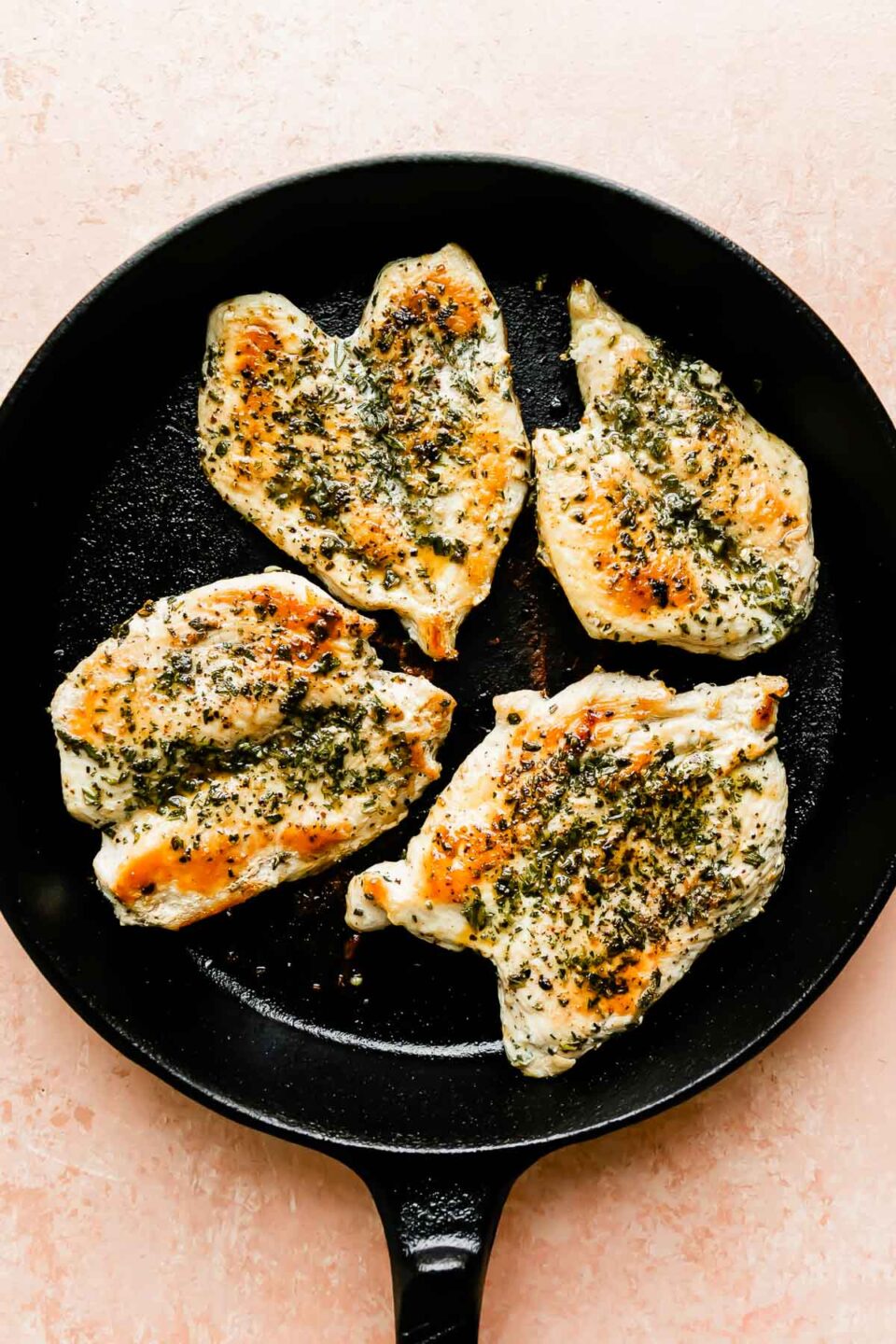 A cast-iron skillet with browned chicken breasts rests atop a pink marbled surface.