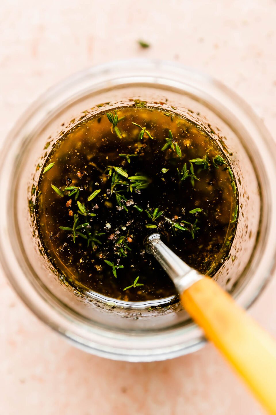 An overhead macro shot of a jar of maple balsamic vinaigrette sitting on a light pink surface. Herbs are scattered in the background.