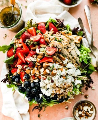 An overhead shot of the strawberry chicken salad on a light pink surface. The bowl is surrounded by a white dish towel, a jar of vinaigrette, and small bowls of strawberries, almonds and goat cheese.