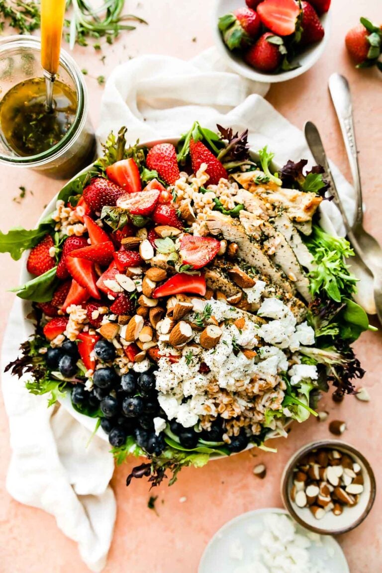 An overhead shot of the strawberry chicken salad on a light pink surface. The bowl is surrounded by a white dish towel, a jar of vinaigrette, and small bowls of strawberries, almonds and goat cheese.