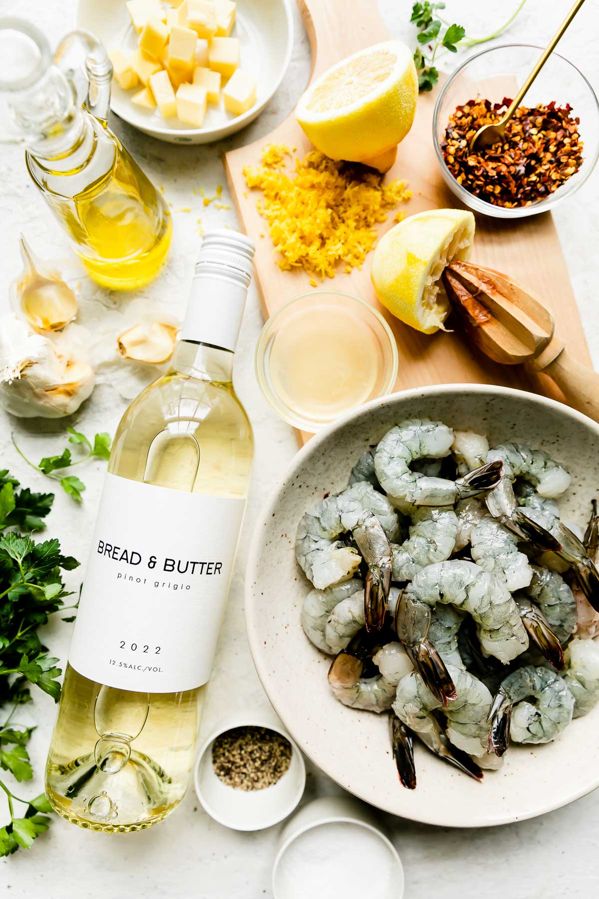 Baked shrimp scampi ingredients arranged on a creamy white textured surface: "extra jumbo" shrimp, olive oil, Bread & Butter California Pinot Grigio or another unoaked dry white wine, garlic, crushed red pepper flakes, unsalted butter, lemon, fresh parsley, kosher salt, and ground black pepper.