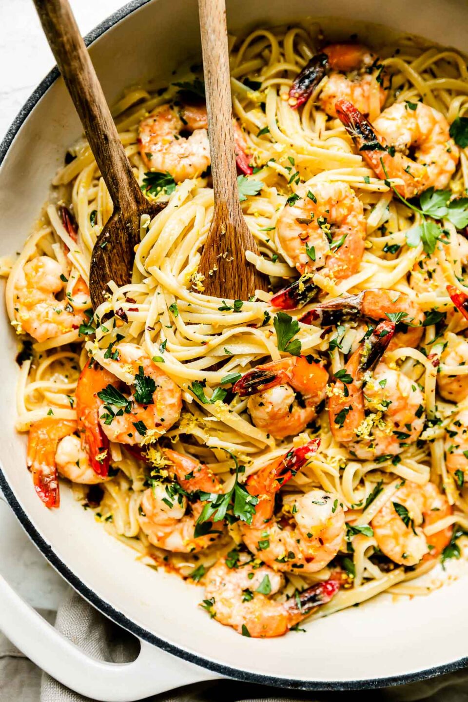 Baked shrimp scampi pasta fills a large double handle braising pan. The pan sits atop a creamy white textured surface. Two wooden serving spoons rest inside of the pan. A cream colored linen napkin rests underneath the pan.