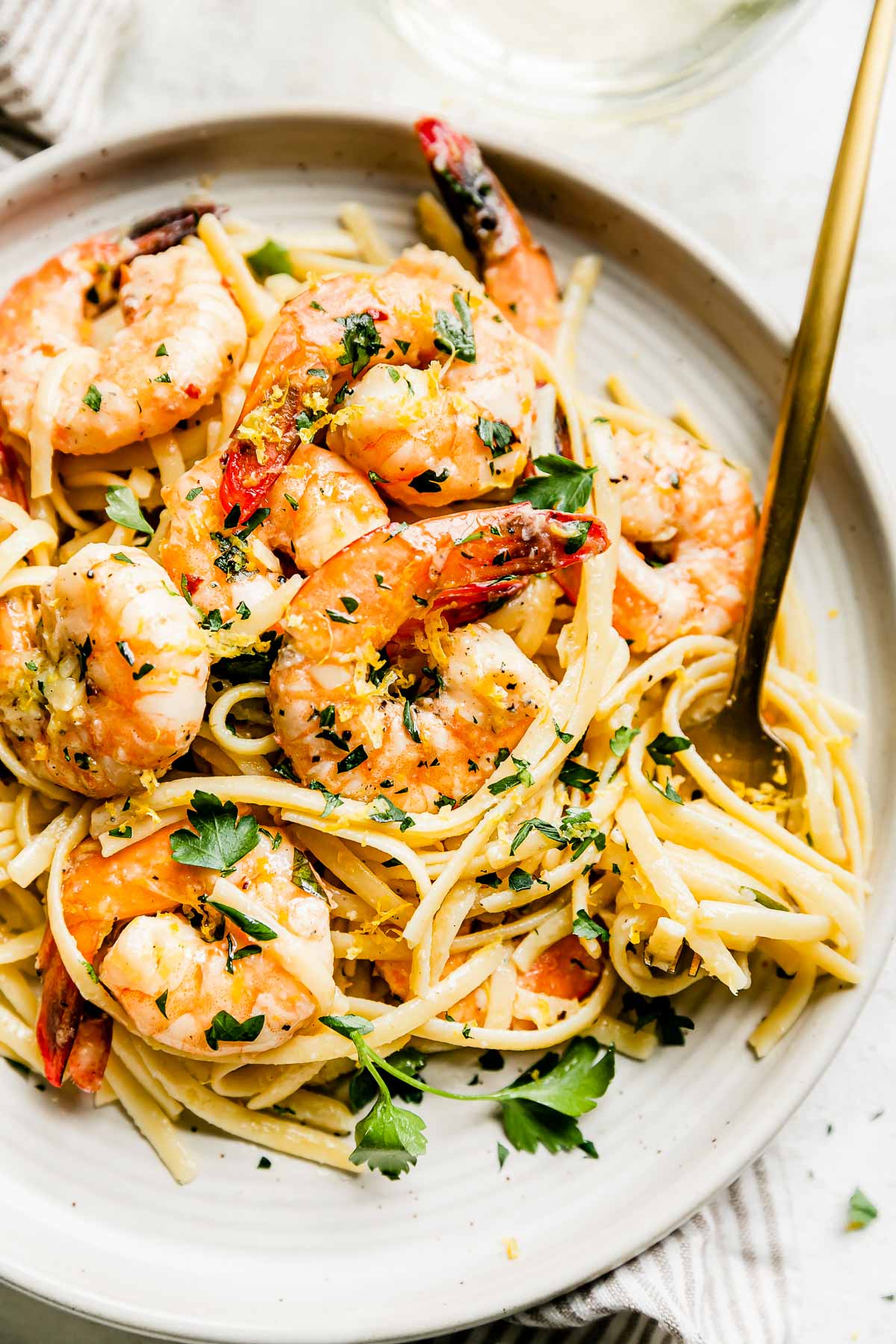 Baked shrimp scampi pasta is served atop a ceramic dinner plate that rests atop a creamy white textured surface. A cream and white striped napkin rests underneath the plate with a gold fork resting atop the plate. A glass of white wine is served alongside the plate of pasta.