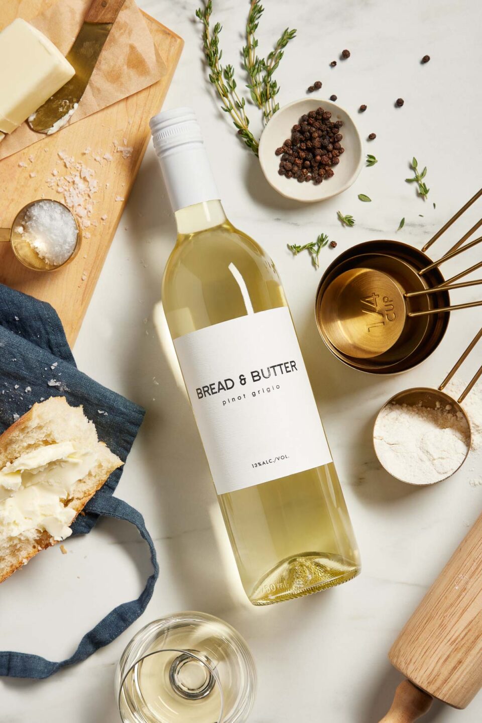 A bottle of Bread & Butter California Pinot Grigio lays flat atop a creamy white textured surface with the label facing upwards. It's surrounded by measuring cups & small bowls filled with ingredients like flour, peppercorns, & fresh herbs.