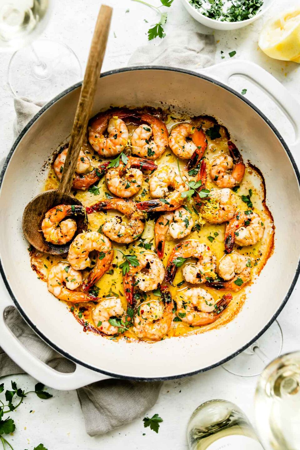 Baked shrimp scampi fills a large double handle braising pan. The pan sits atop a creamy white textured surface. A wooden serving spoon rests inside of the pan and the pan is surrounded by a stack of ceramic plates, two glasses of white wine, a bottle of Bread & Butter California Pinot Grigio, a small white bowl filled with finely chopped fresh parsley, and a spent lemon half. A cream colored linen napkin rests underneath the pan.