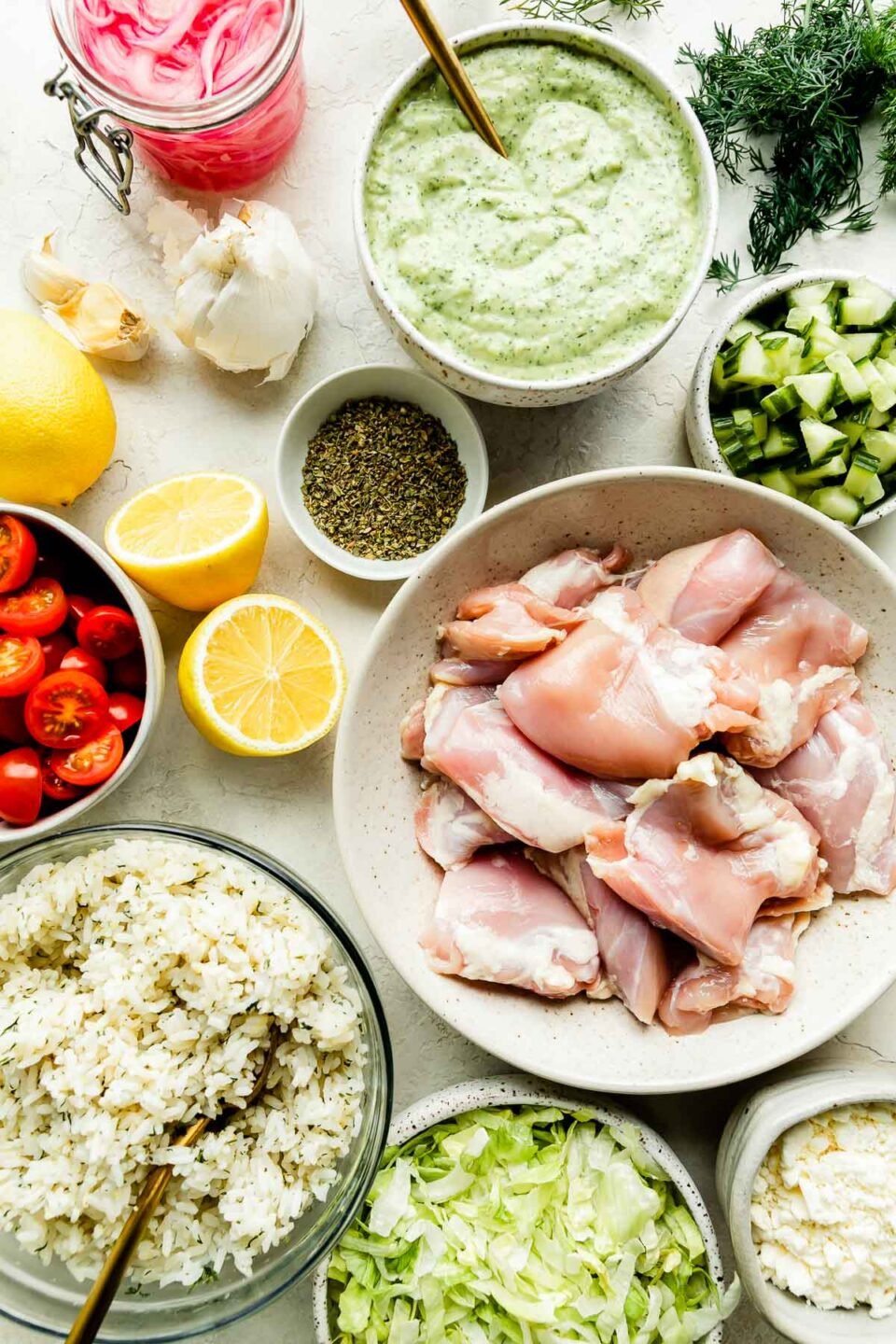 Greek chicken bowl ingredients are displayed on a white marbled surface: chicken thighs, lemons, garlic, dill, oregano, English cucumber, avocado tzatziki, tomatoes, rice, lettuce, feta, & pickled onions.