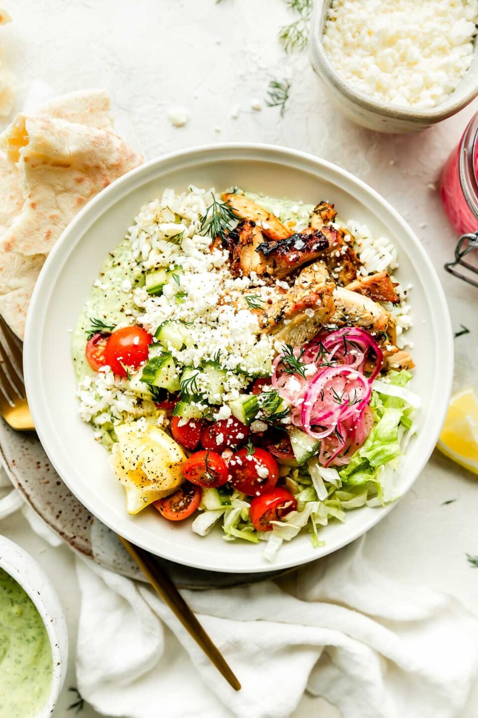 An assembled greek chicken bowl sits on a small plate atop a white surface with a white cloth. A pita and gold fork rest alongside the bowl, and a bowl of avocado tzatziki is on the counter beside it. A bowl of feta, jar of pickled onions, fresh dill, & lemon can be seen in the background.