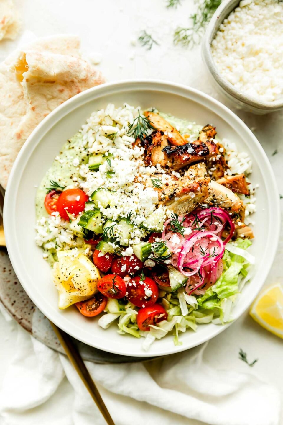 An assembled greek chicken bowl sits on a small plate atop a white surface with a white cloth. A pita and gold fork rest alongside the bowl, and a bowl of avocado tzatziki is on the counter beside it. A bowl of feta, jar of pickled onions, fresh dill, & lemon can be seen in the background.