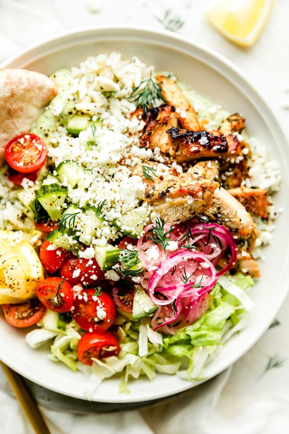 A close-up overhead shot of an assembled greek chicken bowl sitting on a small plate atop a white surface with a white cloth.