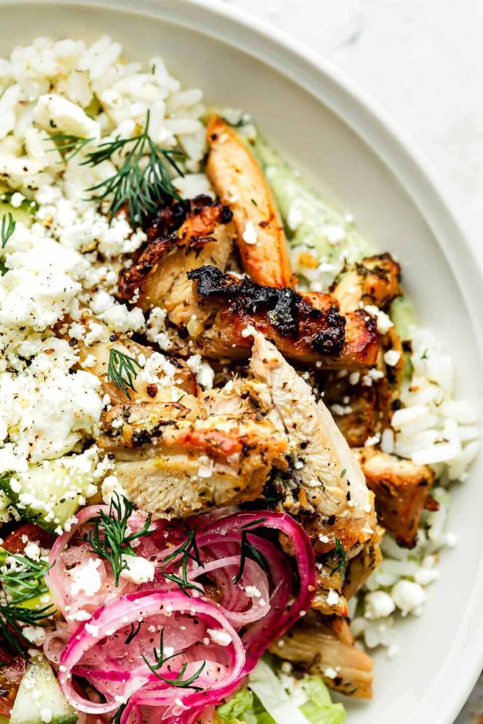 A close-up overhead shot of an assembled greek chicken bowl: rice, chicken, feta, pickled onions, lettuce & diced cucumber.