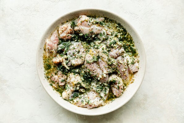 Chicken thighs marinate in a lemon herb marinate in a large white bowl resting on a white textured surface.