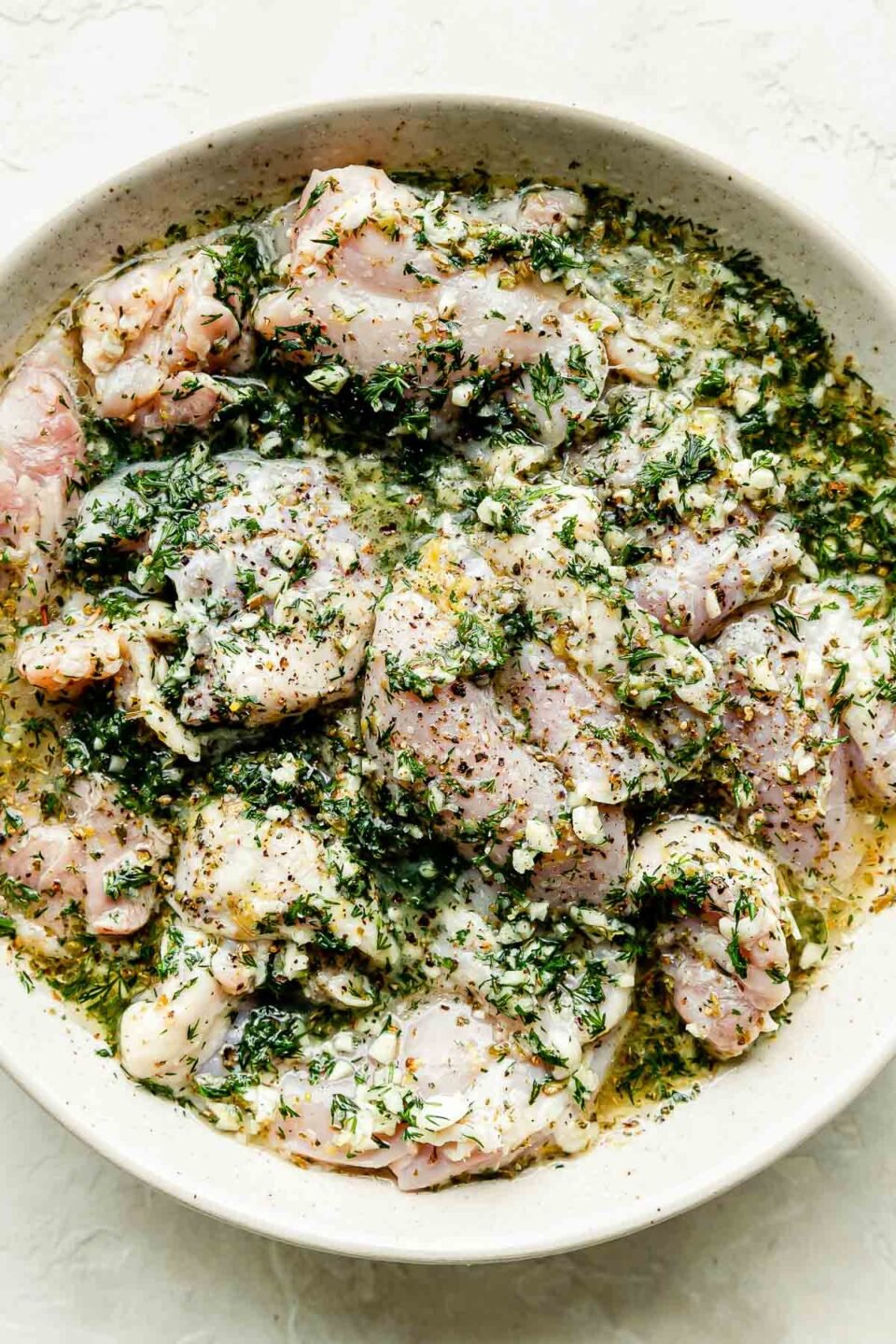 Chicken thighs marinate in a lemon herb marinate in a large white bowl resting on a white textured surface.