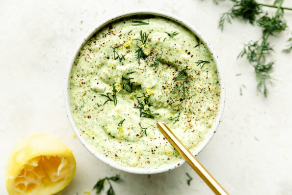 A white bowl of avocado tzatziki sits on a white surface. The tzatziki is topped with fresh dill, and has a gold spoon resting in it. A lemon and fresh dill sit alongside the bowl.