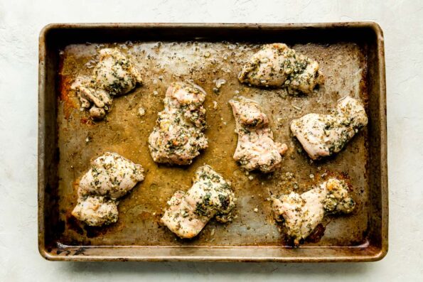Marinated chicken thighs sit on a sheet pan atop a white textured surface.