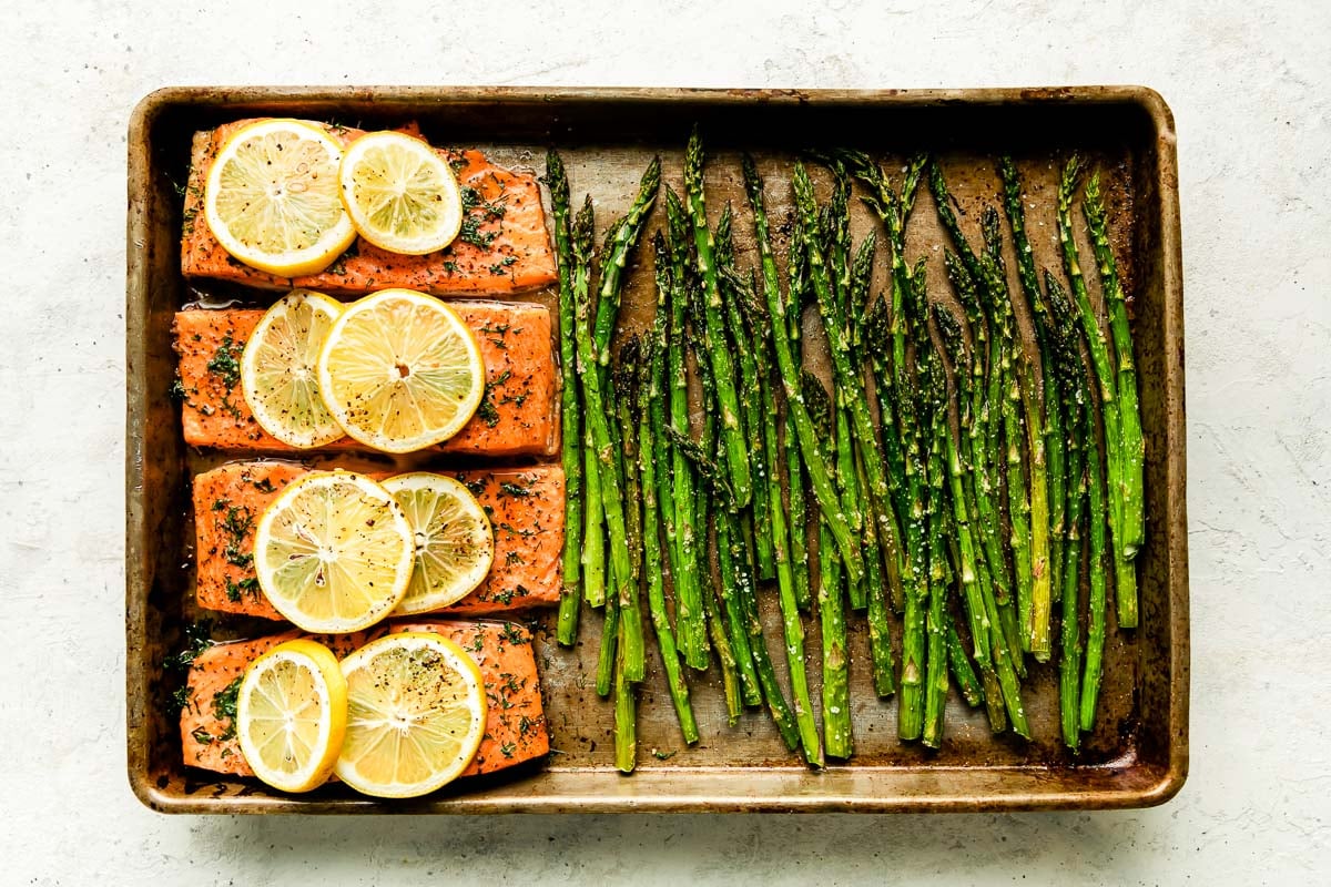 A baked salmon and asparagus sheet pan dinner fills a small metal baking sheet and sits atop a creamy white textured surface.