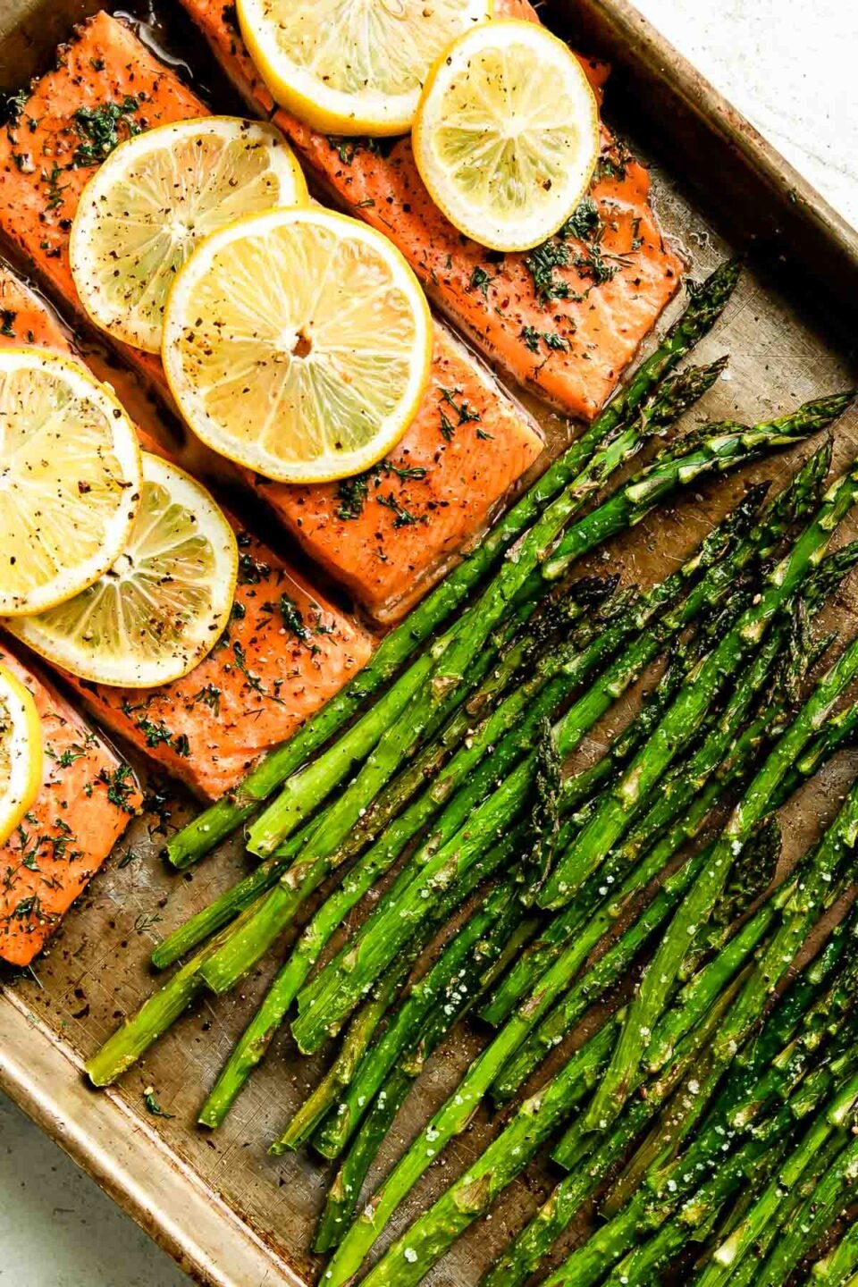 An overhead and close up of baked salmon and asparagus sheet pan dinner fills a small metal baking sheet and sits atop a creamy white textured surface.