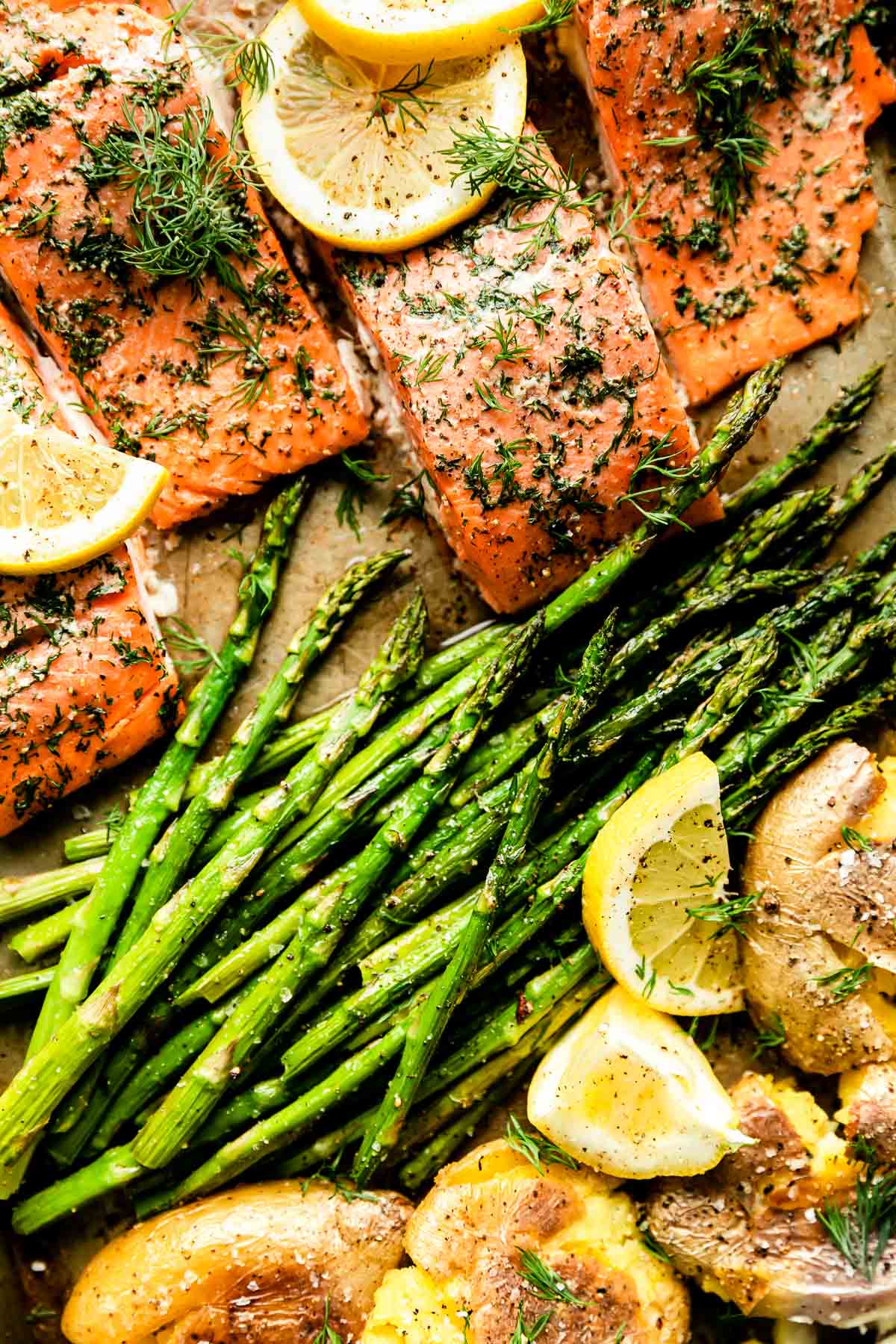 A macro close up shot of baked salmon fillets, asparagus spears, and smashed Yukon gold potatoes arranged on a single sheet pan.
