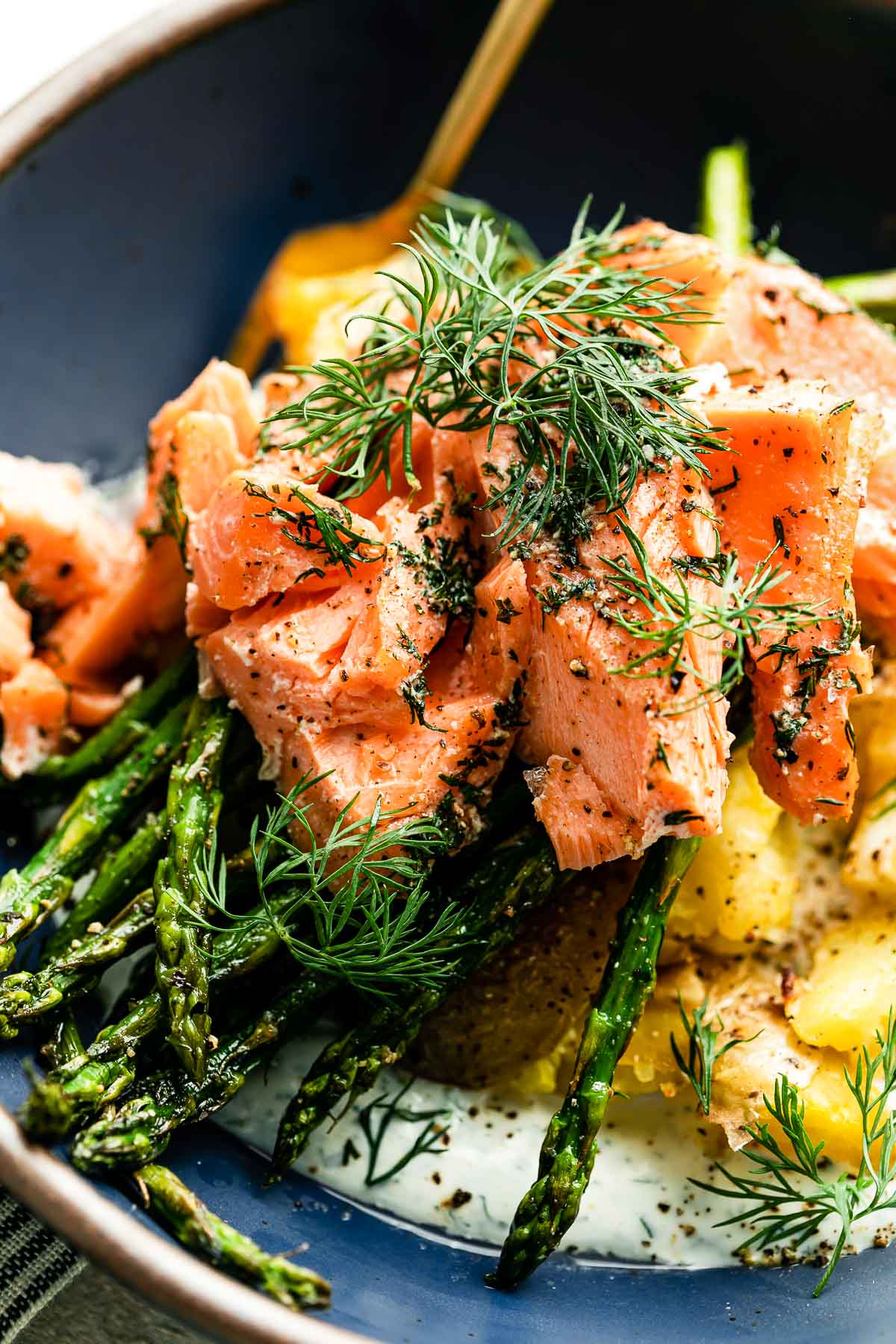 Sheet pan salmon and asparagus with crispy smashed potatoes are served in a blue ceramic bowl with a gold fork and topped with fresh ground pepper and fresh dill.