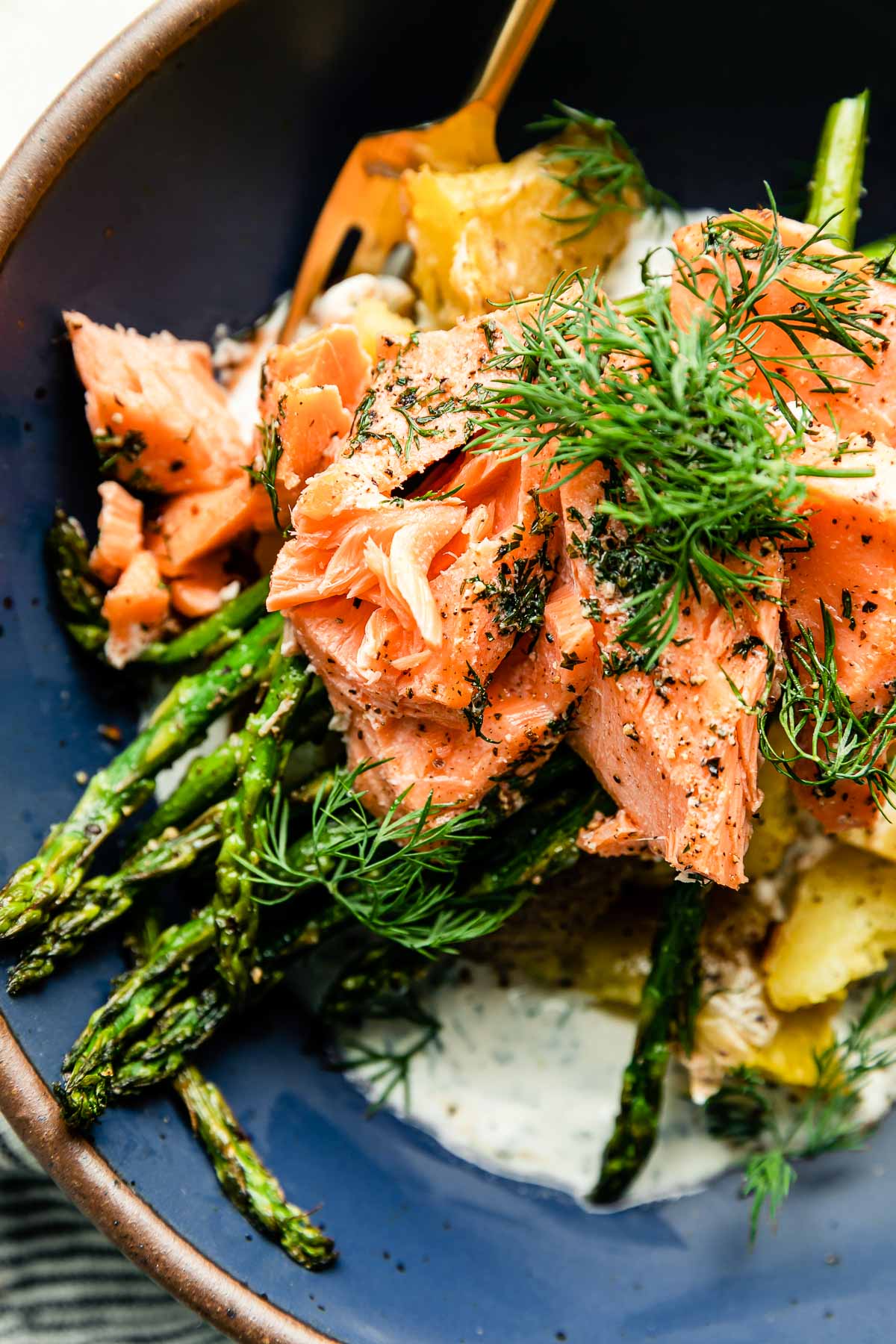 An overhead and macro close up shot of a serving of sheet pan salmon and asparagus served in a blue ceramic bowl that sits atop a creamy white textured surface.