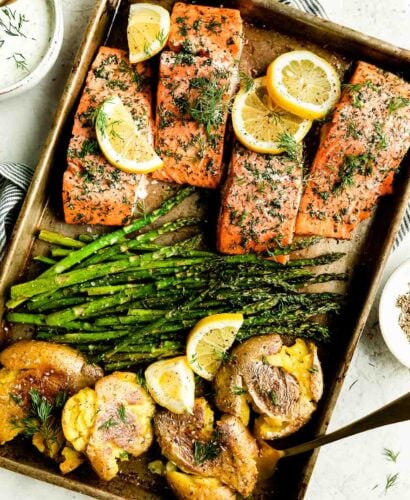 An overhead shot of sheet pan salmon and asparagus with smashed potatoes fills a large metal sheet pan that sits atop a creamy white textured surface. The baking sheet pan is surrounded by a small white ceramic bowl filled with lemon dill yogurt sauce, a blue and white stripped linen napkin, and a small white plate of ground black pepper.