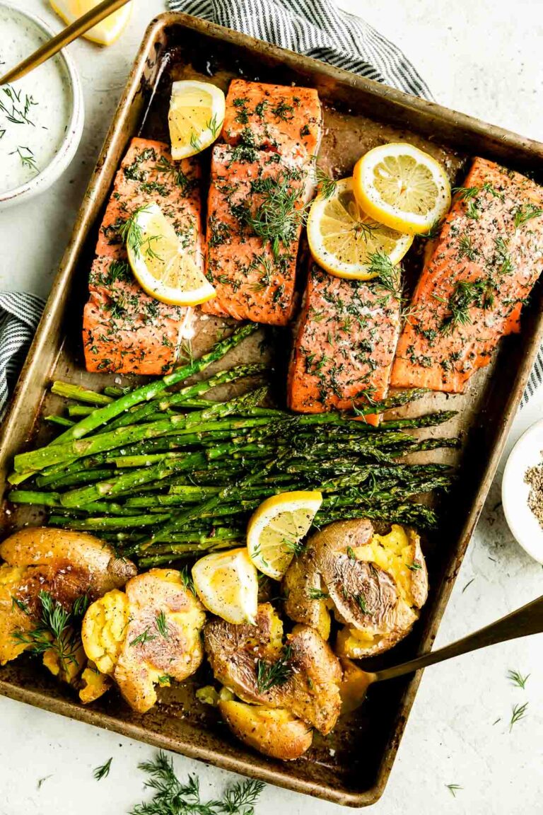 An overhead shot of sheet pan salmon and asparagus with smashed potatoes fills a large metal sheet pan that sits atop a creamy white textured surface. The baking sheet pan is surrounded by a small white ceramic bowl filled with lemon dill yogurt sauce, a blue and white stripped linen napkin, and a small white plate of ground black pepper.