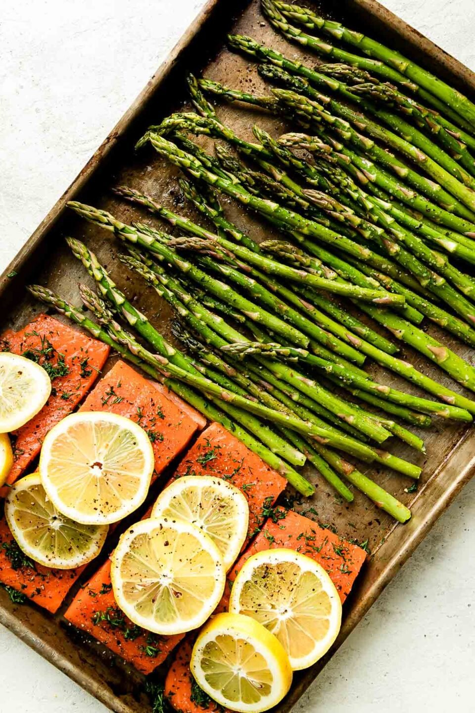 Four seasoned salmon fillets are arranged on a small metal sheet pan topped with lemon. Seasoned asparagus spears are arranged parallel to the fillet and the baking sheet sits atop a creamy white surface.