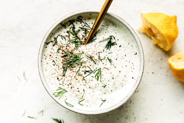 A small white ceramic bowl filled with lemon dill yogurt sauce sits atop a creamy white textured surface. A gold spoon rests inside of the bowl garnished with fresh dill and ground black pepper. Two lemon wedges rest alongside the bowl.