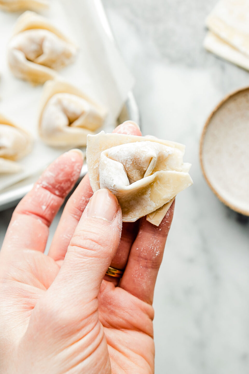 An overhead shot of a hand holding a folded wonton. A tray of prepared wontons, extra wrappers & a dish of water can be seen in the background on a white marbled surface.