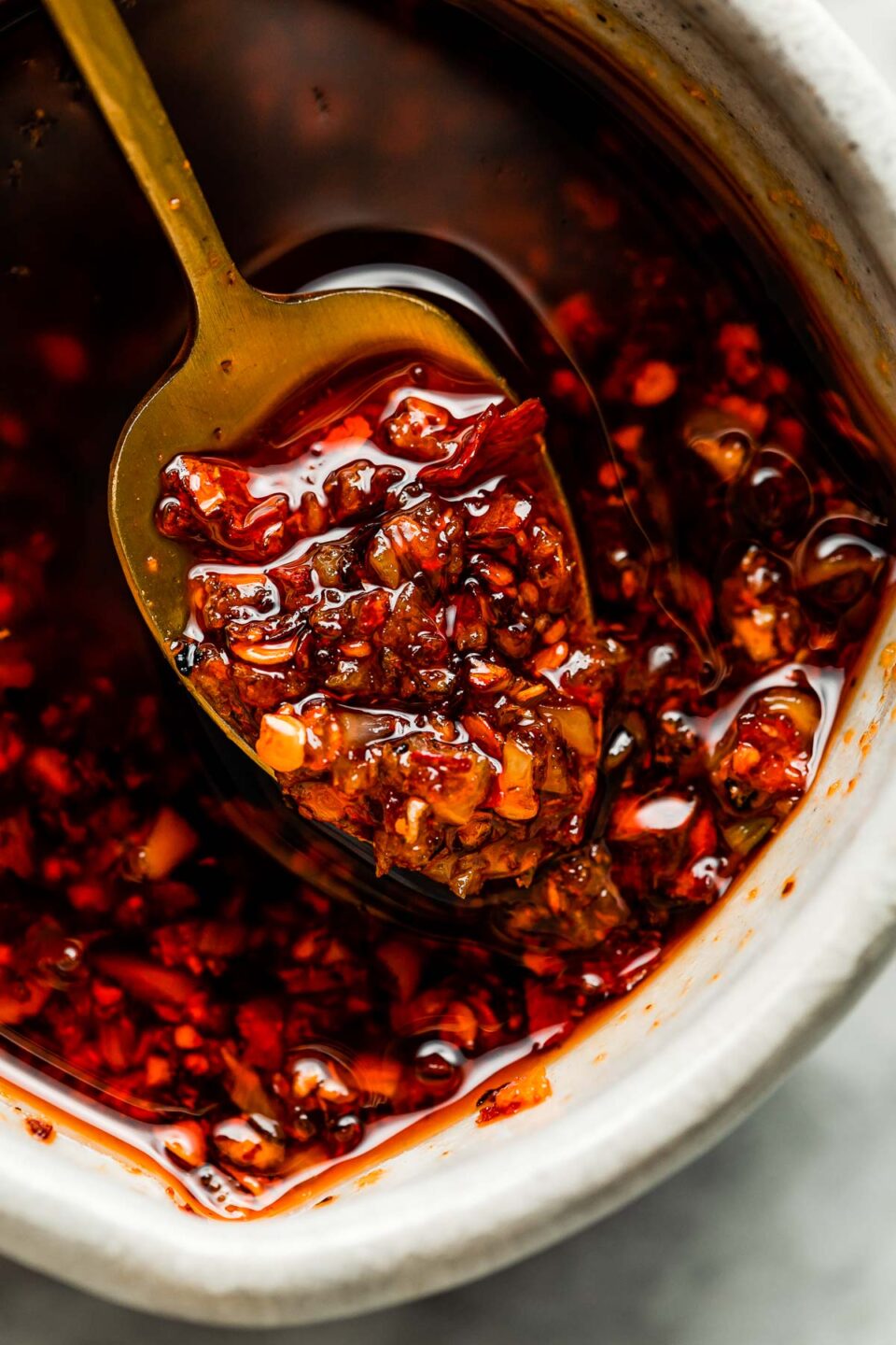 A close-up macro shot of a spoon of spicy chili oil being held up over a small bowl of spicy chili oil.