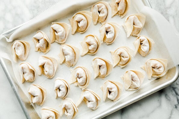 A parchment-paper lined sheet pan full of prepared wontons sits atop a white marbled surface.