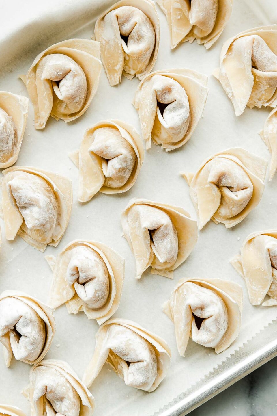 An overhead shot of parchment-paper lined sheet pan full of prepared wontons sitting atop a white marbled surface.