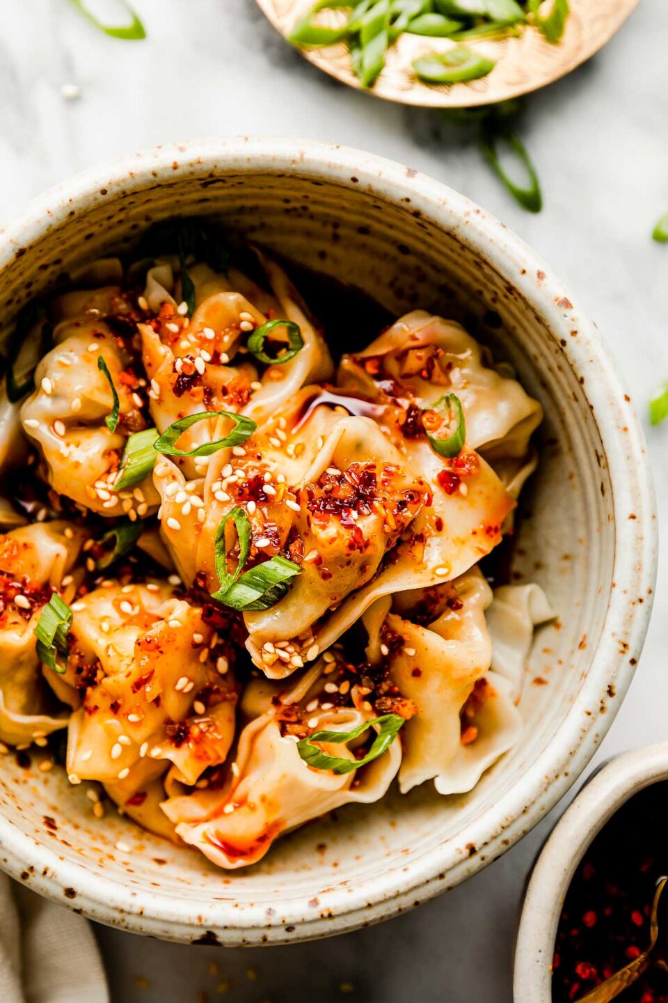 An overhead shot of a stoneware bowl of cooked wontons, topped with spicy chili oil and sliced green onions atop a white marbled surface. Smaller dishes of sliced green onions & spicy chili oil sit beside it.