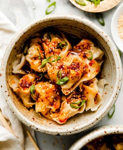 An overhead shot of a stoneware bowl of cooked wontons, topped with spicy chili oil and sliced green onions atop a white marbled surface alongside chopsticks and a white dish towel. Smaller dishes of sliced green onions, spicy chili oil, and sesame seeds sit beside it.